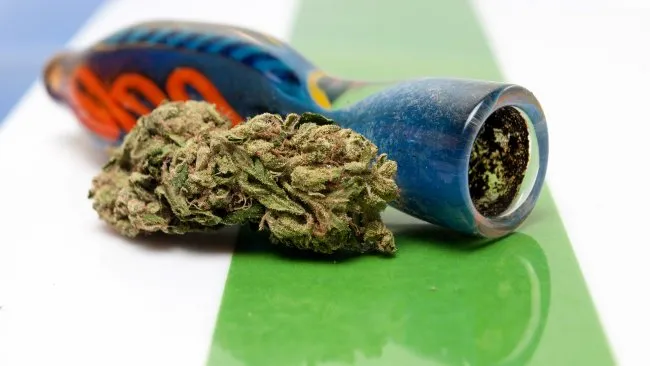 a small chillum cannabis pipe next to a large bud of cannabis, on a green and white surface
