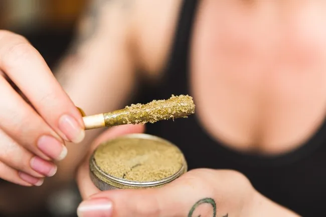 woman holding a joint covered in cannabis kief in one hand and a silver metal circle disc that is holding tan colored powder