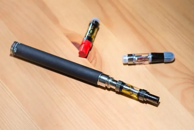 a black vape pen battery with a cartridge attached, with 2 cartridges next to it, on a wooden background