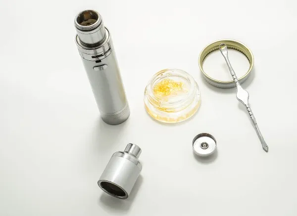 a silver dab pen taken apart next to a container of cannabis wax and a metal dab tool